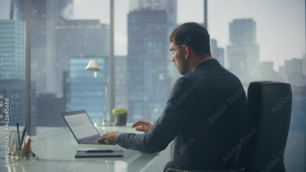 Relaxed Businessman in a Suit Sitting at a Desk in Modern Office, Using Laptop Computer, Next to Window with Big City with Skyscrapers View. Successful Finance Manager Planning Work Projects. Relaxed Businessman in a Suit Sitting at a Desk in Modern Office, Using Laptop Computer, Next to Window with Big City with Skyscrapers View. Successful Finance Manager Planning Work Projects.