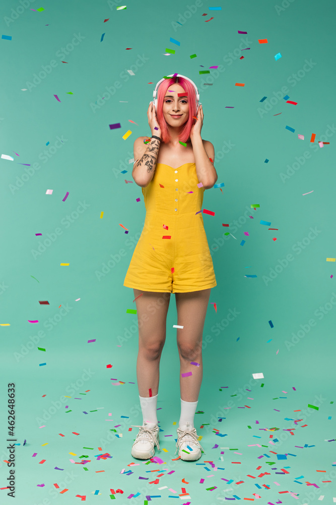 full length of happy young woman with pink dyed hair in wireless headphones near colorful confetti on blue full length of happy young woman with pink dyed hair in wireless headphones near colorful confetti on blue