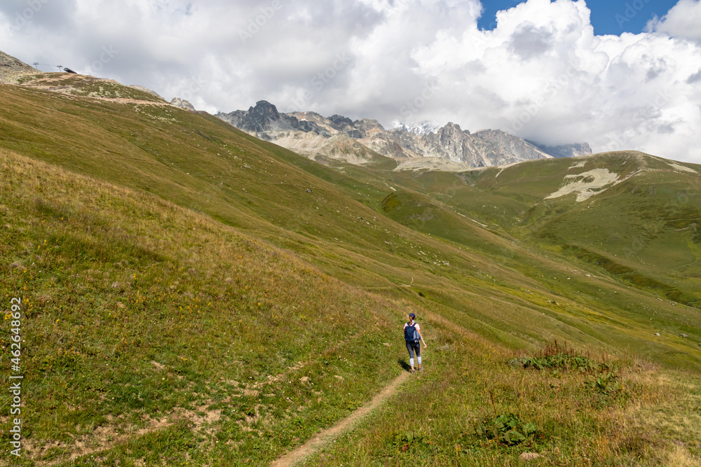 A woman hiking along a narrow pathway in high Caucasus mountains in Georgia. There are high glaciers in the back. Thick clouds above the sharp peaks. Lush pastures on the sides. Barren peaks.