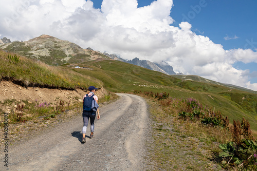 A woman hiking along a gravelled road in high Caucasus mountains in Georgia. The road leads to a ski resort on a steep slope. Thick clouds in the back. Lush pastures on the sides. Barren peaks.