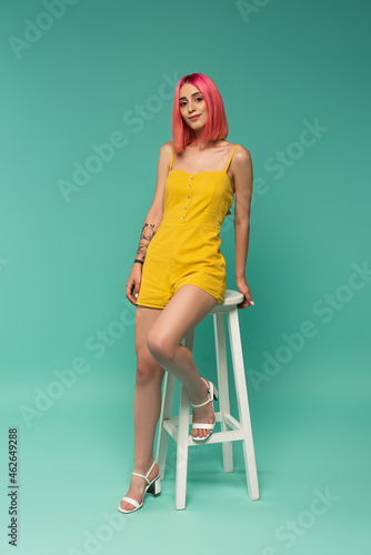 full length of happy young woman with pink dyed hair sitting on stool on blue