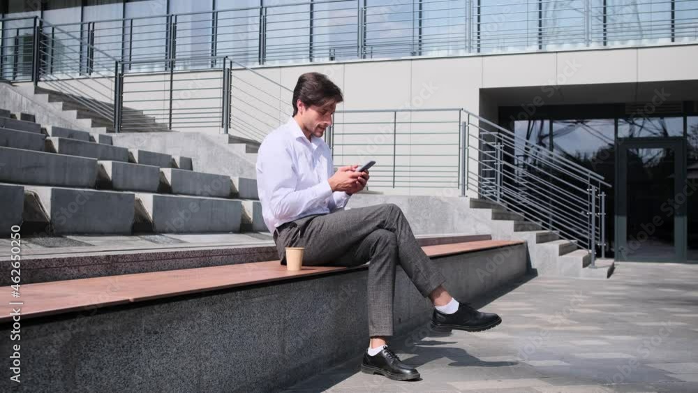 Businessman using mobile phone and drinking coffee sitting on bench against office building Businessman using mobile phone and drinking coffee sitting on bench against office building