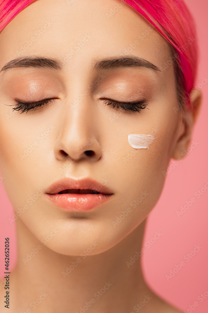 close up of young woman with closed eyes and face cream on cheek isolated on pink