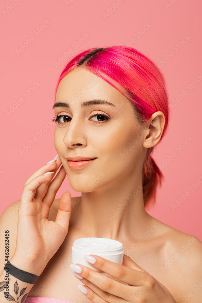 tattooed young woman with dyed hair smiling while holding container and applying face cream isolated on pink tattooed young woman with dyed hair smiling while holding container and applying face cream isolated on pink