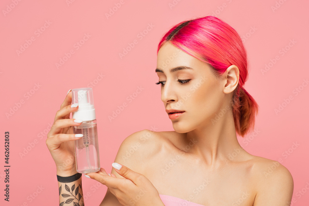 tattooed young woman with colorful hair looking at bottle with toner isolated on pink tattooed young woman with colorful hair looking at bottle with toner isolated on pink