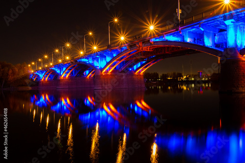 Blue and golden lights of illuminated Communal bridge over the Yenisei River in Krasnoyarsk, Russia. Reflection of a bridge and illuminations in the water. Night urban landscape. City Light