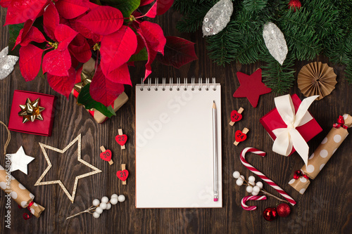 Empty white notebook, red flower poinsettia, Christmas decorations, gift boxes and candy canes on dark wooden background.