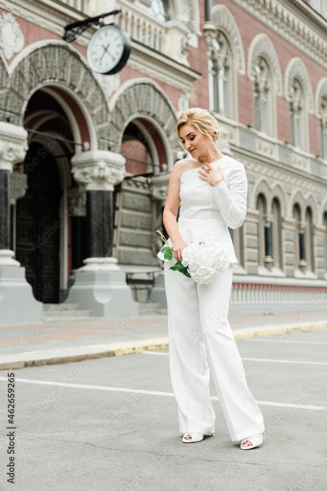 A young stylish lady in a white pantsuit with an elegant hairstyle with a bouquet of hydrangea flowers on the streets of the city. The woman is the bride. Soft selective focus