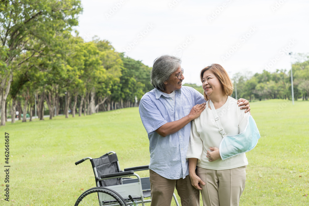 Asian elderly husband takes care his elderly sick wife while sitting on a wheelchair with injured arm wrapped in cast in the park. Asian senior couple spending time and relaxing together outdoor Asian elderly husband takes care his elderly sick wife while sitting on a wheelchair with injured arm wrapped in cast in the park. Asian senior couple spending time and relaxing together outdoor
