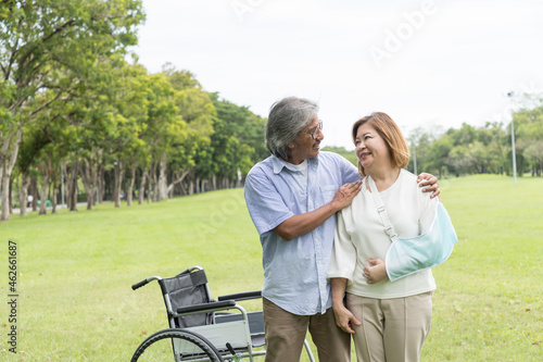 Asian elderly husband takes care his elderly sick wife while sitting on a wheelchair with injured arm wrapped in cast in the park. Asian senior couple spending time and relaxing together outdoor