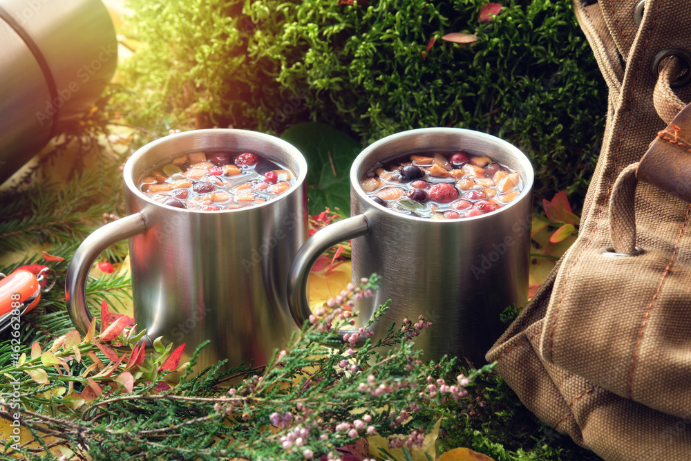 Hiking mugs of healthy fruit tea, canvas backpack of traveller, thermos, fallen autumn leaves, forest moss, sprigs of heather and spruce in autumn forest. - obrazy, fototapety, plakaty 