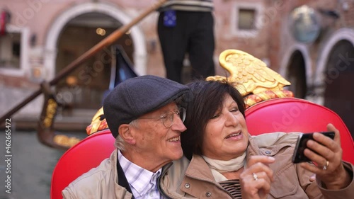 Beautiful romantic mature married couple rides on a gondola boat and takes a video call or selfies in Venice, in autumn fall, or winter. Happy holidays in Europe, Italy.