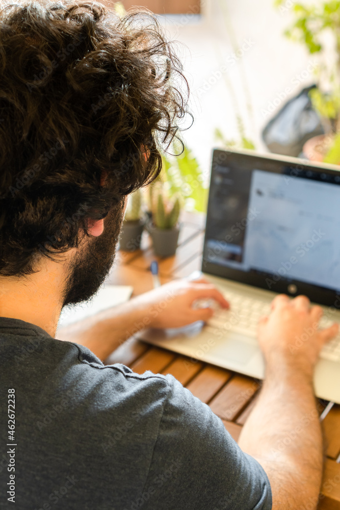 Back view of young man with beard working with a laptop in a sunny outdoor place full of plants at home.