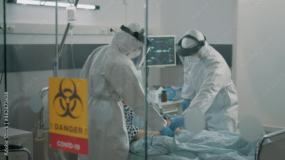 Medical staff in hazmat suits consulting aged patient with coronavirus. Sick woman in hospital ward quarantine receiving checkup visit from paramedics with protective uniforms and masks Medical staff in hazmat suits consulting aged patient with coronavirus. Sick woman in hospital ward quarantine receiving checkup visit from paramedics with protective uniforms and masks