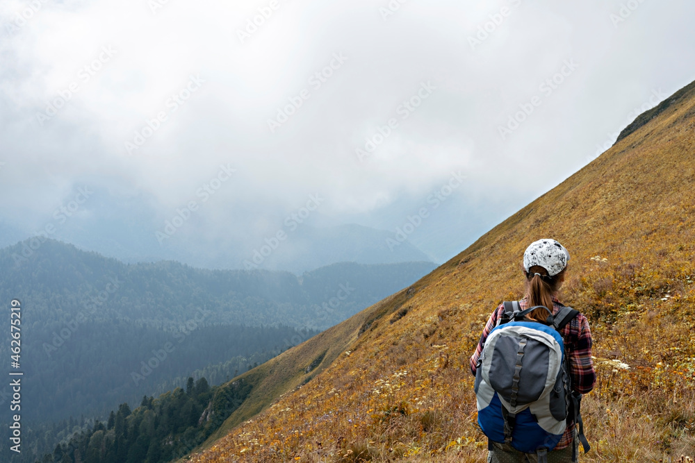 rear view of young woman hiker in cap with large hiking backpack looking at the mountain view of the Aibga ridge of the Caucasus mountains, healthy active lifestyle, weekend activities, landscape
