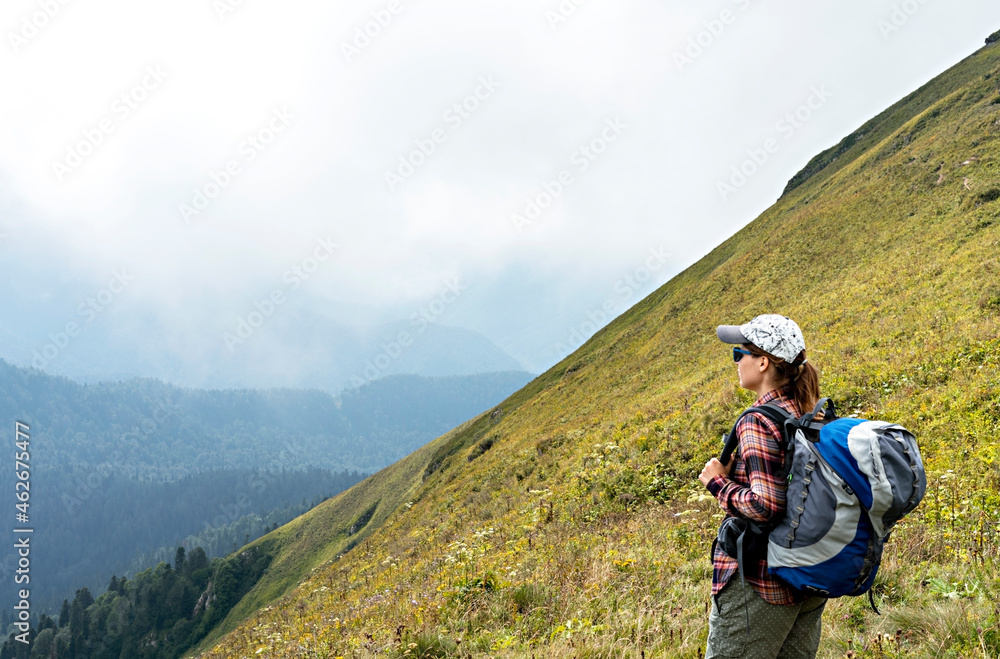 Young woman hiker in cap and sunglasses with large hiking backpack looking at mountain view of the Aibga ridge of the Caucasus mountains, healthy active lifestyle, weekend activities beauty in nature