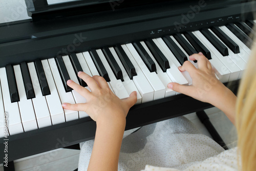 A blonde little baby girl is playing a digital piano in the living room, close-up from the back. A child learns to play a musical instrument. Early music education for children