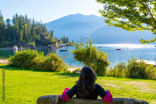 A young brunette woman seen from behind as she relaxes on a park bench at Kaslo Bay Park along Kootenay Lake in Kaslo, BC, Canada. photo