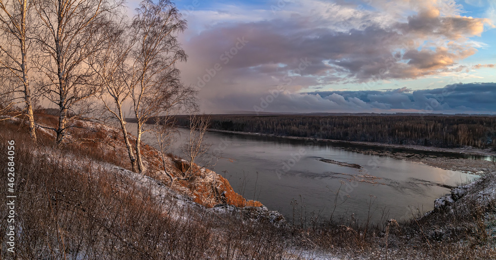 colorful autumn river view. first snow on the hills, frozen river, mountains, white birch trees on the slopes and pink sunset clouds, stormy sky