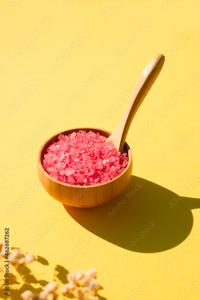 Wooden bowl with a spoon filled with pink bath sea salt. Beauty treatment for spa and wellness on yellow background. Wooden bowl with a spoon filled with pink bath sea salt. Beauty treatment for spa and wellness on yellow background.