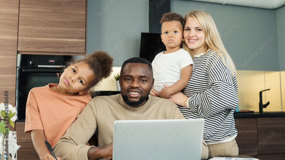Portrait of beautiful loving diverse family of four looking at camera and smiling indoors. Black entrepreneur man working on laptop from home surrounded by White wife and two biracial kids Portrait of beautiful loving diverse family of four looking at camera and smiling indoors. Black entrepreneur man working on laptop from home surrounded by White wife and two biracial kids