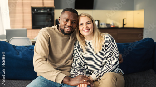 Fotografia Portrait of beautiful diverse couple of Black man and Caucasian woman looking at camera and smiling sitting on sofa at home