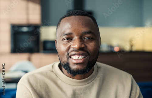 Head and shoulders portrait of handsome young Black man looking at camera and smiling happily sitting on sofa. Happy guy with beard posing at home