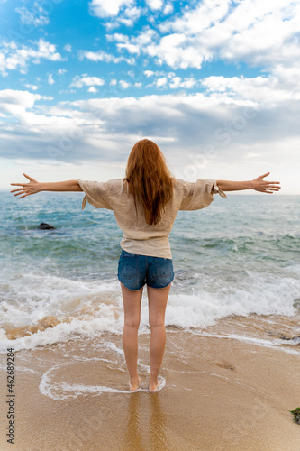 Back view of redheaded young woman standing at seafront with arms outstretched