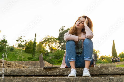 Young redheaded tired woman sitting on steps next to a laptop in a park photo