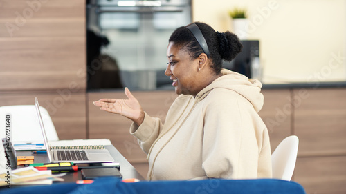 Fotografie Side view of smiling Black plump woman having video call on laptop and gesturing sitting at desk working from home