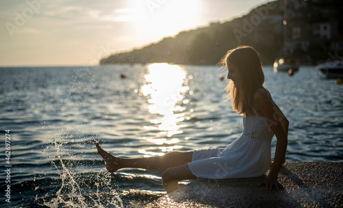 Croatia, Lokva Rogoznica, girl sitting at waterside during sunset splashing with water