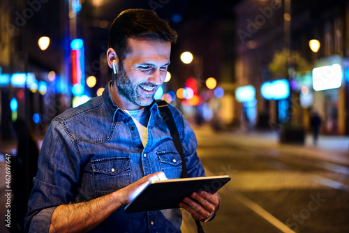 Smiling man with wireless headphones using digital tablet in the city at night
