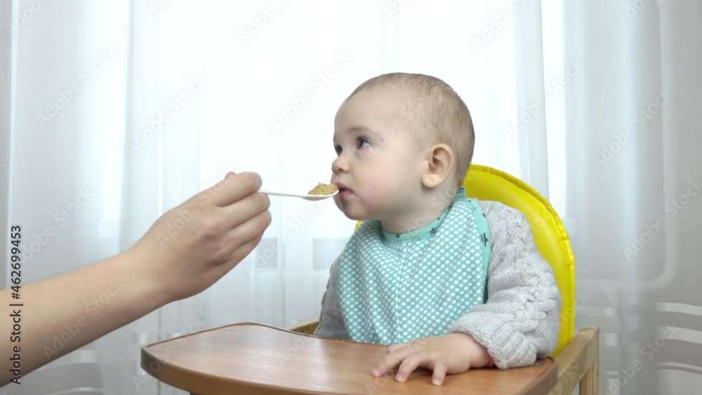 Mom feeds the milky girl baby from a spoon on a highchair Mom feeds the milky girl baby from a spoon on a highchair
