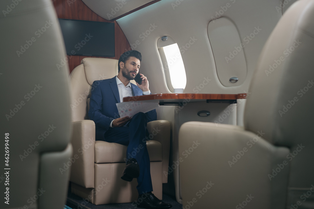 Handsome pensive middle eastern businessman talking on mobile phone, holding financial report working with documents sitting in plane. Confident entrepreneur flying private jet, successful business Handsome pensive middle eastern businessman talking on mobile phone, holding financial report working with documents sitting in plane. Confident entrepreneur flying private jet, successful business