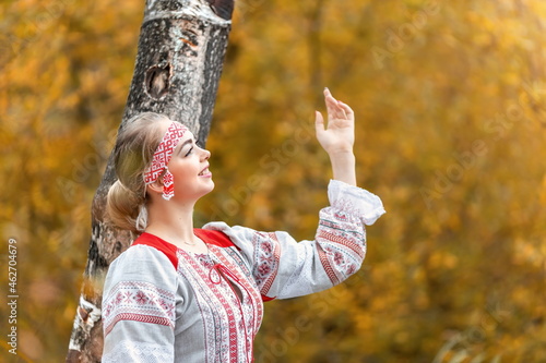 Portrait of young beautiful smiling woman in folk traditional slavic clothes greets the sun at nature in autumn forest.