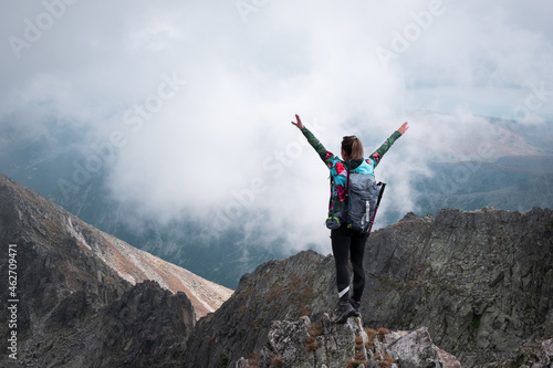 Hiker girl standing on the edge of a rocky, steep cliff with hands in the air, backpack and walking sticks on a misty, foggy day