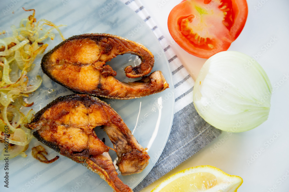 Fried fish with onions lies on a plate, on a waffle towel, next to a lemon and a tomato on a white background. High quality photo Fried fish with onions lies on a plate, on a waffle towel, next to a lemon and a tomato on a white background. High quality photo