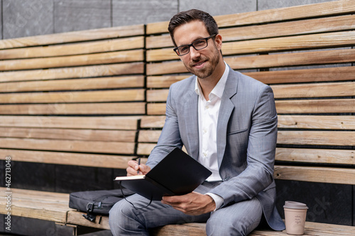 Businessman sitting on wooden bench in the city writing into notebook