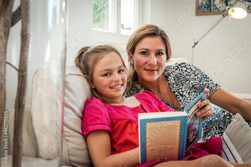 Portrait of smiling mother and daughter with book on couch in living room photo