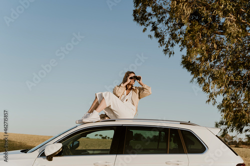Female traveller looking through binoculars sitting on white car roof