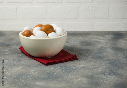 Eggs in a white ceramic bowl on a red napkin on the gray surface of the table. White brick wall. Copy space for text.