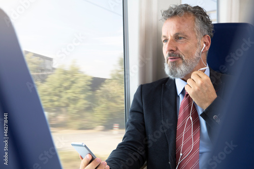 Mature businessman sitting train, using earphones and smartphone