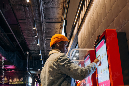 Smiling African-American man in warm denim jacket with wireless earphones uses self-service kiosk to order snack in cafe