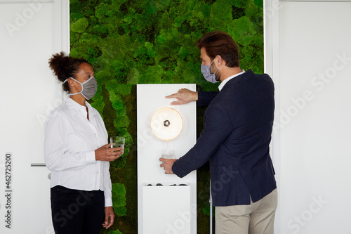 Colleagues with protective masks keeping distance at water dispenser during Corona crisis