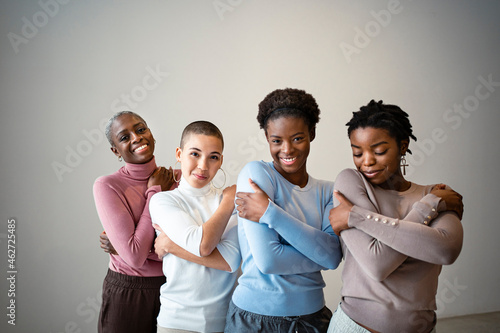 Smiling female friends self hugging against wall at home photo