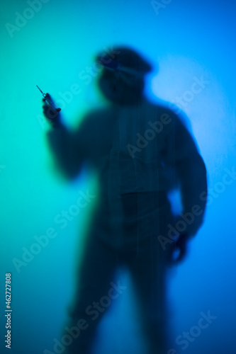 Woman in protective wear behind translucent glasspane holding syringe photo