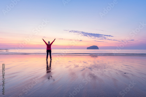 Mature woman with arms raised standing at Seacliff Beach during sunset, North Berwick, Scotland