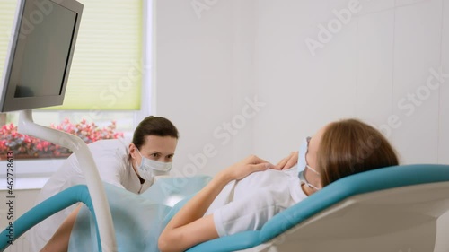 Experienced female gynecologist in lab coat, face mask and gloves holding medical vaginal speculum for examining patient. Young woman lying on gynecological chair during check up. photo