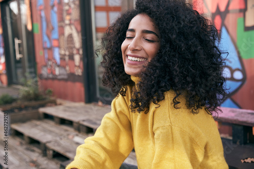 Smiling young woman with eyes closed sitting against wall in city photo