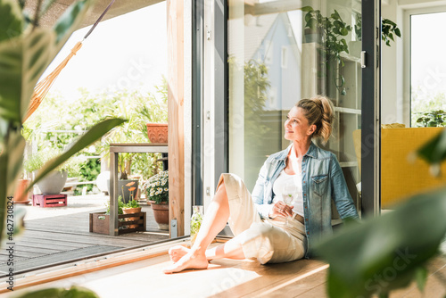 Smiling mature woman sitting on the floor at open terrace door looking at distance photo